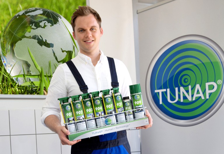 Qualität & Service Junger Mechaniker hält einen mit Aerosol-Dosen gefüllten Werkstatt-Korb und auf der Hintergrundwand ist eine gläserne Weltkugle und ein TUNAP-Logo sichtbar